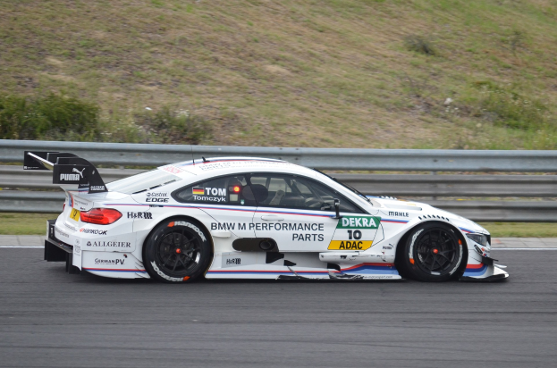 BMW M4 DTM-Rennwagen auf einer Rennstrecke mit Sponsorentext, umgeben von grünem Gras und einer Begrenzung im Hintergrund.
