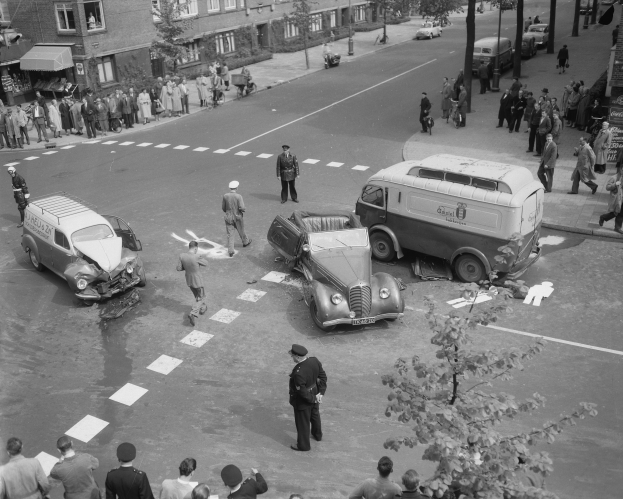 Schwarzes und weißes Bild eines Autounfalls an einer Straßenkreuzung mit mehreren Fahrzeugen, darunter ein Lieferwagen, und einer Gruppe von Zuschauern; Gebäude, Bäume, Pfosten und Texttafeln sind im Hintergrund zu sehen.