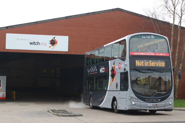 Doppeldeckerbus mit Rauchwolken an einem Gebäude vorbeifahrend, mit Gras, Bäumen und einem blauen Himmel im Hintergrund und einem sichtbaren Kfz-Kennzeichen am Bus.