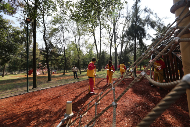 Gruppe von Menschen, die einen Seilparcours in einem Park mit Bäumen im Hintergrund und einer Stange mit Seilen auf der rechten Seite absolvieren.