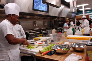Eine Gruppe von Köchen in einer Küche, die Essen auf einem Schneidbrett zubereiten, mit einem Koch im Vordergrund in einer weißen Schürze und Mütze, der ein Messer hält, umgeben von Utensilien, Schüsseln, Gemüse, einem Mobiltelefon, Flaschen und anderen Gegenständen, mit einem Fernseher und Deckenlampen im Hintergrund.