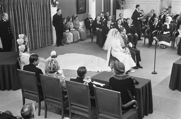 Ein Schwarz-Weiß-Foto einer Hochzeitszeremonie mit Menschen, die sitzen und stehen, einem Mann mit Mikrofon und einer Frau mit Blumenstrauß, mit einem Rednerpult, Blumengestecken, einem Vorhang, einem gerahmten Foto an der Wand und einer Tür im Hintergrund.