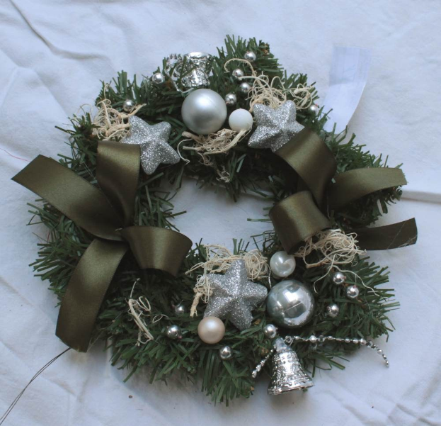 Ein festlicher Adventskranz mit silbernen Schmuckelementen und Bändern, der bunte Kugeln, Sterne und weitere dekorative Gegenstände enthält und auf einem weißen Tuch liegt.