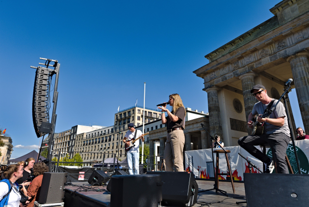 Eine Gruppe von Menschen, die auf einer Bühne vor dem Brandenburger Tor in Berlin Musik spielen, umgeben von Lautsprechern und Equipment, unter einem klaren blauen Himmel.