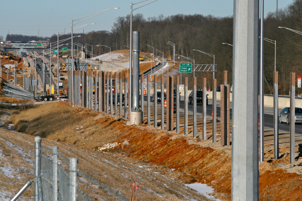 Ein belebter Baustelle mit verschiedenen Fahrzeugen, Masten, Lichtern und Texttafeln, umgeben von einem Zaun, Gras, Schnee und Büschen unter einem Himmel.
