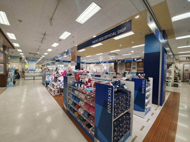 Das Innere eines offiziellen Tokyo 2020-Shops mit Kleiderstangen, Namensschildern mit Text, Deckenlampen und verschiedenen anderen Gegenständen.