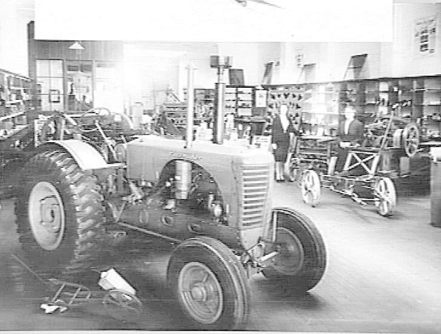 Ein Schwarz-Weiß-Foto eines Traktors in einer Fabrik mit mehreren Personen drumherum, anderen Fahrzeugen auf dem Boden, einem Müllimer auf der rechten Seite und Regalen mit diversen Gegenständen im Hintergrund.