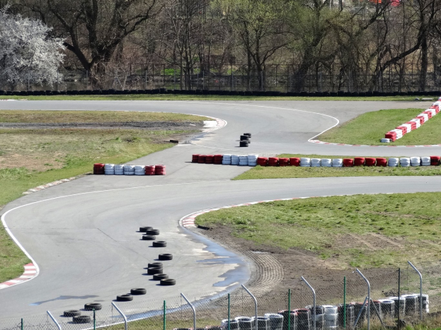 Rennstrecke mit zahlreichen Reifen entlang ihrer Ränder, gesäumt von Gras und Maschendrahtzaun, mit im Hintergrund sichtbaren Bäumen.