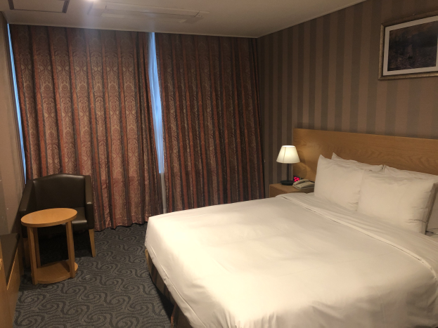 Hotelzimmer mit einem großen Bett, Stuhl, Tisch, Telefon, Lampe, Foto auf dem Wand, Fenster mit Vorhängen und Deckenbeleuchtung.