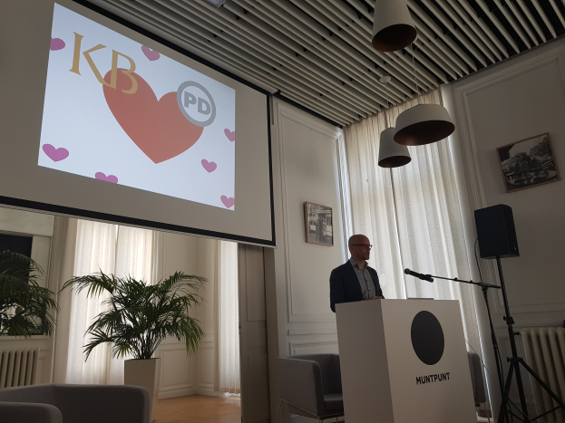 Ein Mann steht an einem Rednerpult mit einem Mikrofon und spricht zu einem Publikum in einem Raum mit einer Projektionswand, Stühlen, einer Pflanze, Fotorahmen, Vorhängen und Deckenleuchten, wahrscheinlich über Geschlechtergerechtigkeit.