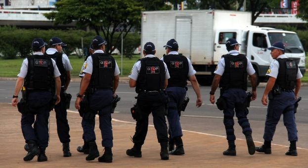 Eine Gruppe von Beamten in Uniform auf einer Straße mit einem geparkten Lkw auf der rechten Seite, Gebäude, Bäume und eine Brücke im Hintergrund.