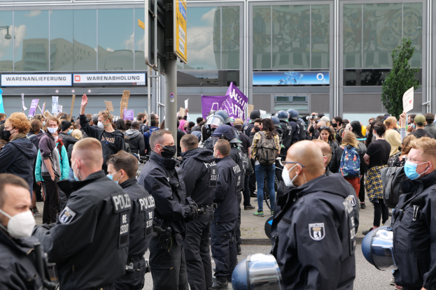 Eine große Gruppe von Menschen steht vor einem Gebäude, einige halten Schilder und tragen Helme, mit einem Pfahl mit einer Schildertafel im Vordergrund und einem Baum im Hintergrund, scheinbar protestierend.