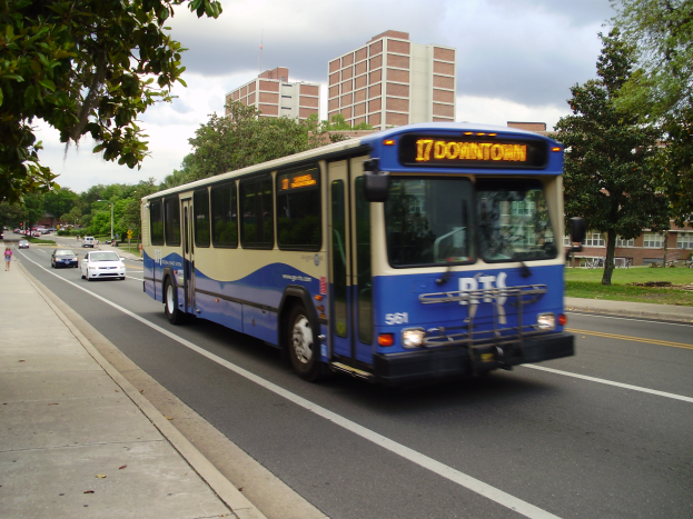 Ein blauer und weißer Shuttlebus fährt eine von Hochhäusern gesäumte Straße entlang, mit einigen Fußgängern auf dem linken Gehweg und Bäumen, Pfählen und einem klaren blauen Himmel im Hintergrund.