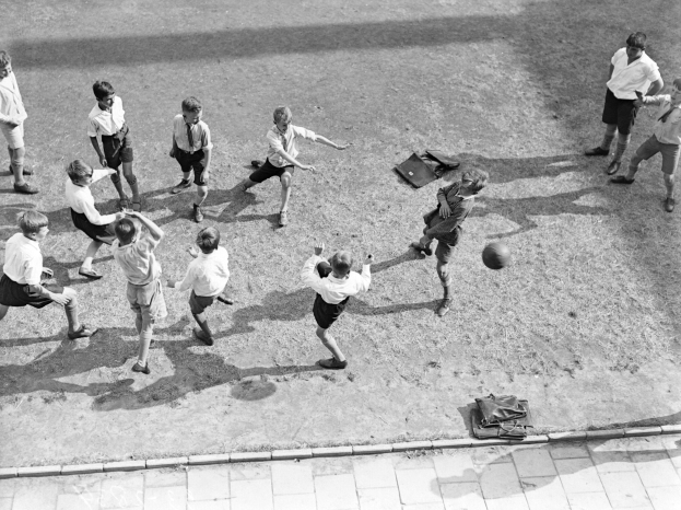 Eine Gruppe junger Jungs, die auf einem Feld Fußball spielen, mit kurzen Hosen und Schuhen, mit Taschen auf dem Boden drumherum, in Schwarz-Weiß.