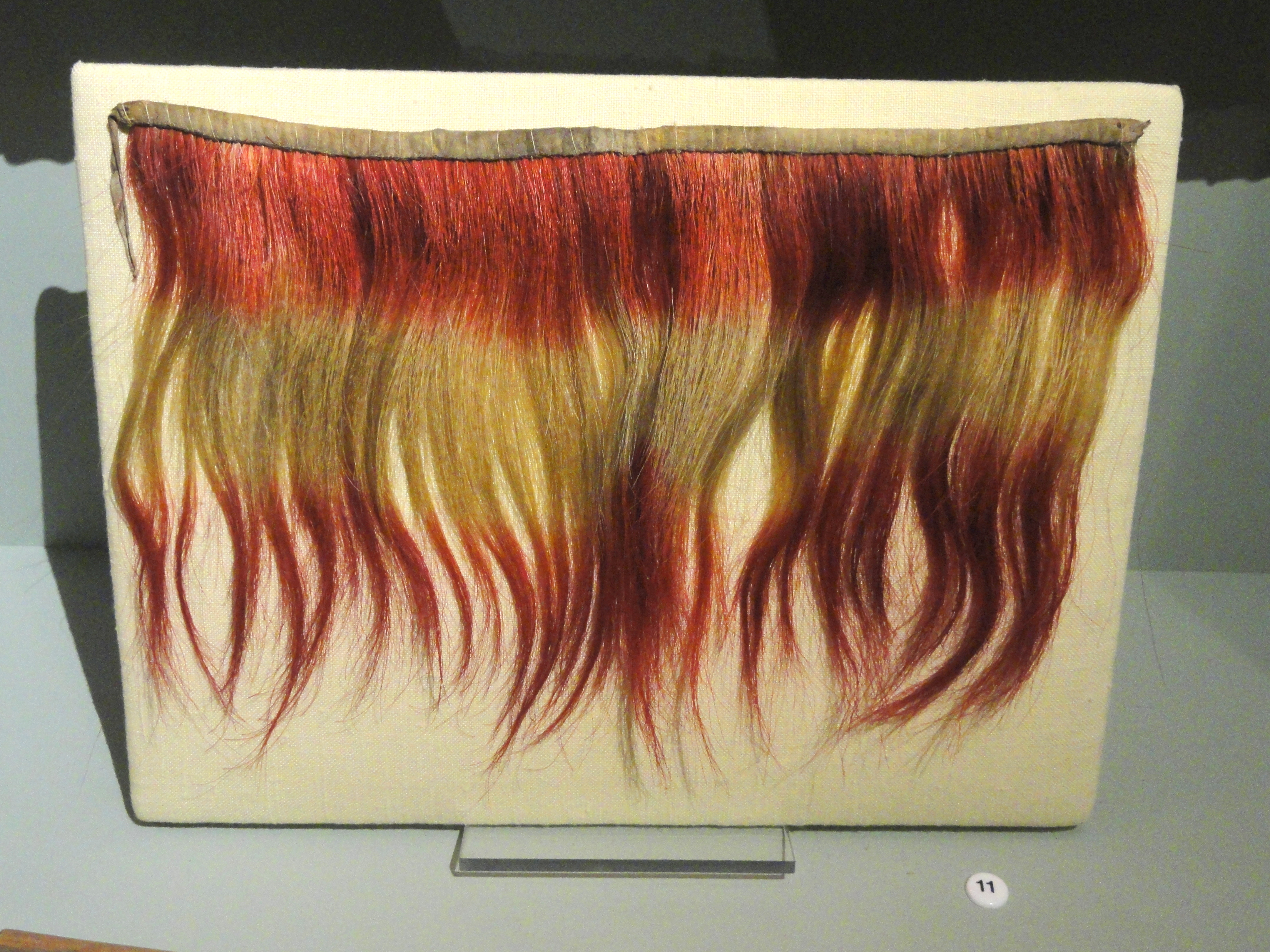 Nahaufnahme einer leuchtenden, bunt gefärbten Haarprothese auf einem Brett in einem Museums Setting.
