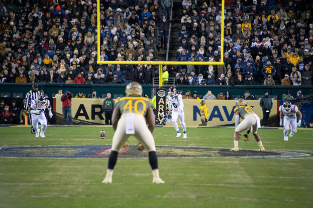 Fußballspieler in Helmen und Uniformen auf einem Feld mit Zuschauern auf den Rängen und Stadionwerbung im Hintergrund.