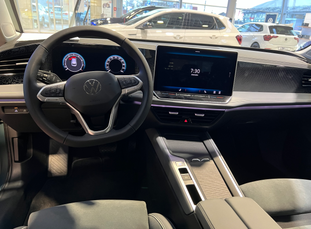 Das Innere eines Volkswagen e-Golf mit einem Lenkrad, Armaturenbrett, Bildschirmen, Knöpfen und Sitzen, mit einer Aussicht auf andere Autos, Gebäude, Bäume, Pfosten und den Himmel durch die Glastüren.