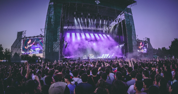 Eine große Menge an einem Musikfestival schaut auf einer hell erleuchteten Bühne mit Instrumenten und Bildschirmen, die vor einem Gebäude, Bäumen und einem klaren blauen Himmel liegen.