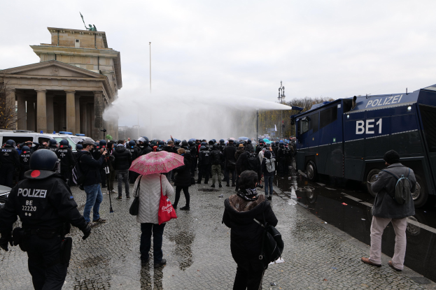 Eine Gruppe von Menschen vor einer Reihe von Polizisten in Helmen und Uniformen, einige mit Schirmen, vor einem Gebäude mit Säulen und Bögen, Fahrzeugen, Bäumen und Laternen in Berlin, Deutschland.
