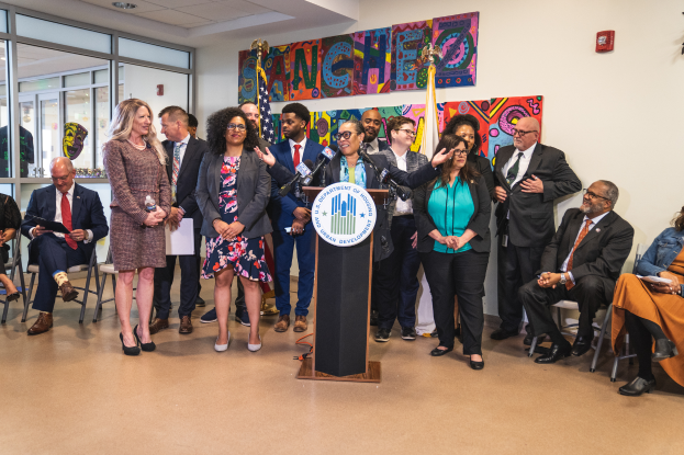 Eine Gruppe von Menschen, die vor einem Podium mit Mikrofon und einer Tafel mit Text stehen und sitzen, mit Flaggen, einer Tafel, einem Feuerlöscher und Deckenleuchten an der Rückwand.
