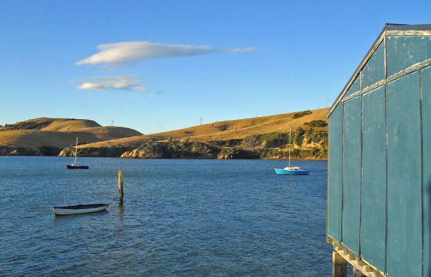 Boote und Masten im Wasser, ein Holzhaus rechts, mit Bäumen und Bergen mit Windmühlen im Hintergrund, unter einem bewölkten Himmel.