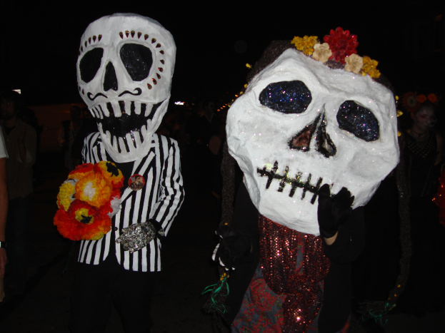 Zwei Personen in buntfarbenen, kunstvoll verzierten Tag-der-Toten-Kostümen, jede mit einem Blumenstrauß in der Hand, vor einem dunklen Hintergrund.