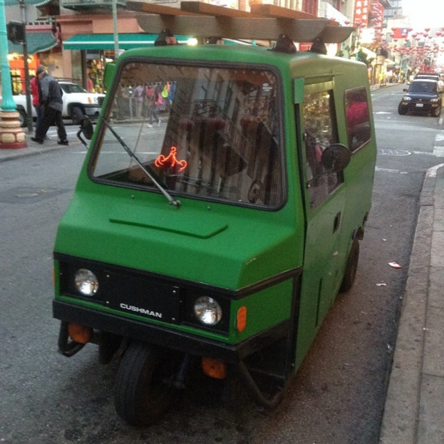 Ein grünes Fahrzeug steht auf einer Straße mit Gebäuden, Masten, anderen Fahrzeugen und einigen Menschen im Hintergrund.