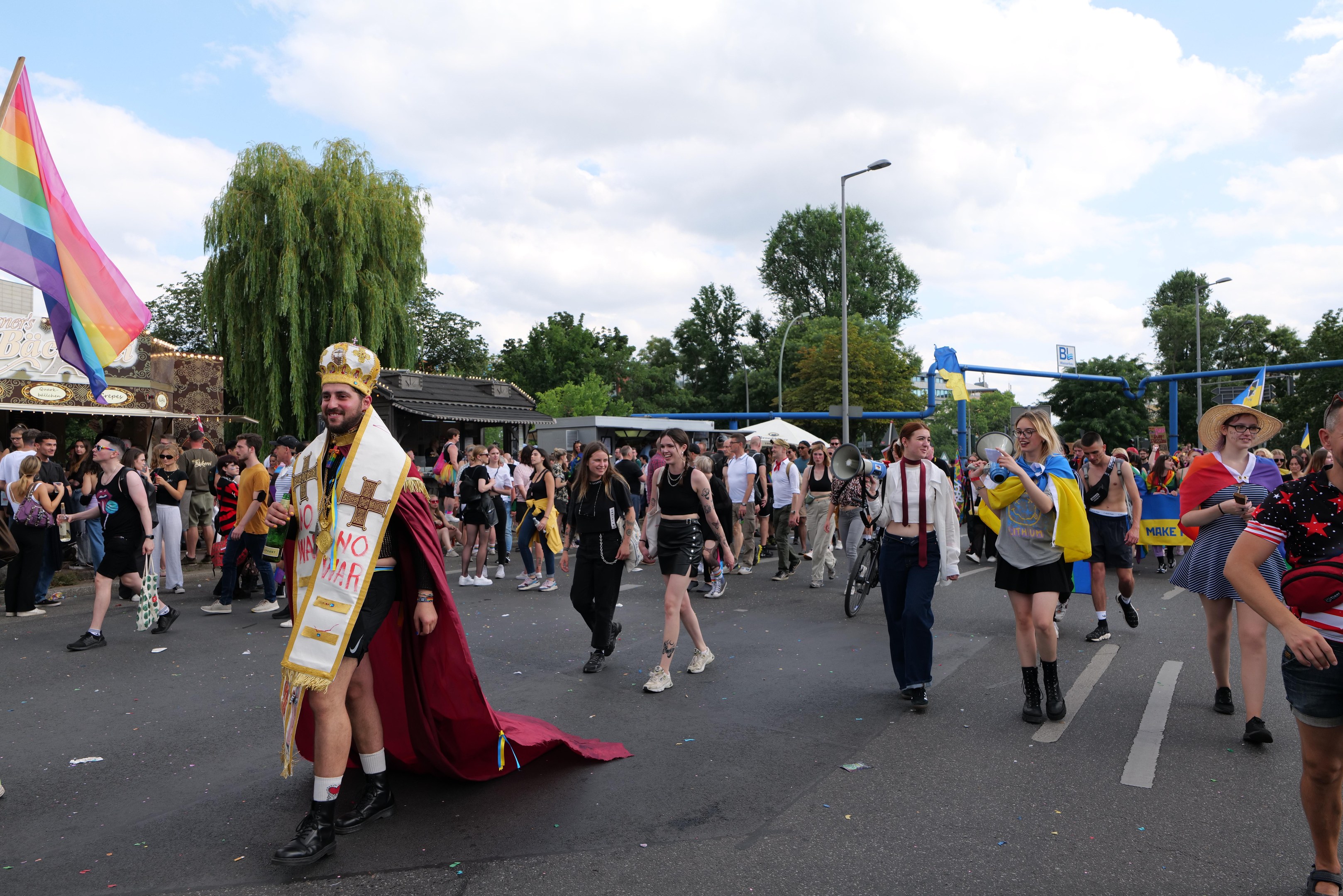 Eine Gruppe von Menschen marschiert bei der Pride Parade 2018 mit einer Regenbogenflagge und Musikinstrumenten, einige tragen Mützen, vor einem Hintergrund aus Laternenmasten, Bäumen, Hütten und einem bewölkten Himmel.