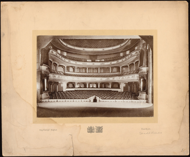 Ein altes Schwarz-Weiß-Foto des Auditoriums der Royal Albert Hall in London, das die Bühne mit Reihen von Stühlen, Säulen, Geländern und Text sowie ein Logo unten auf einem dunklen Hintergrund zeigt.