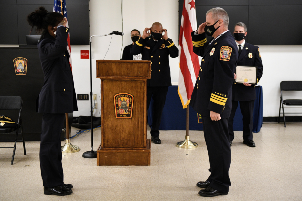 Eine Gruppe uniformierter Personen in Masken steht vor einem Podium mit Mikrofon, umgeben von Flaggen, Stühlen und Tischen, während einer Vereidungszeremonie für einen neuen Feuerwehrchef, wobei eine Person ein Zertifikat hält.