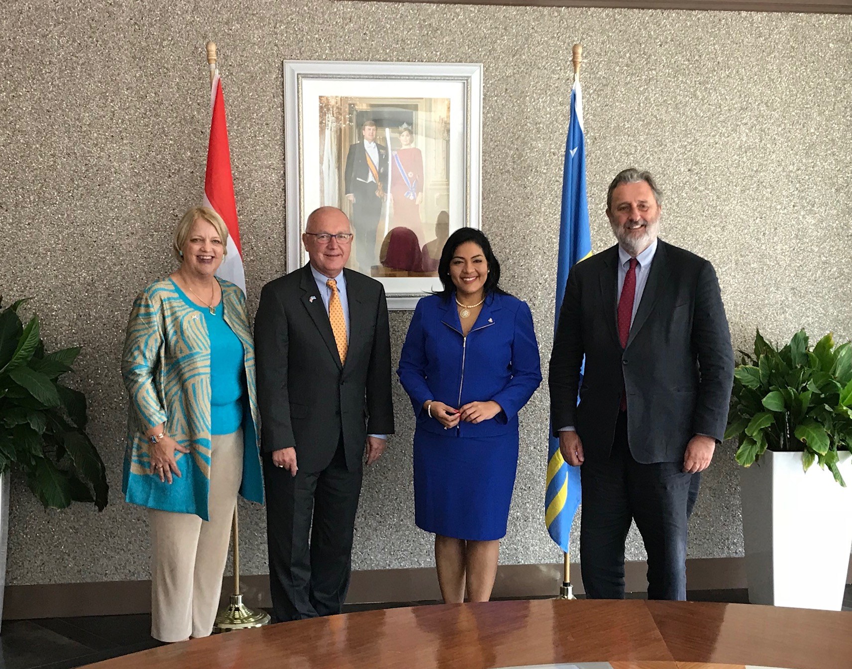 Eine Gruppe von Menschen, die um einen Tisch mit Papieren stehen und von Fahnen, Topfpflanzen und einem Rahmenfoto an der Wand umgeben sind.