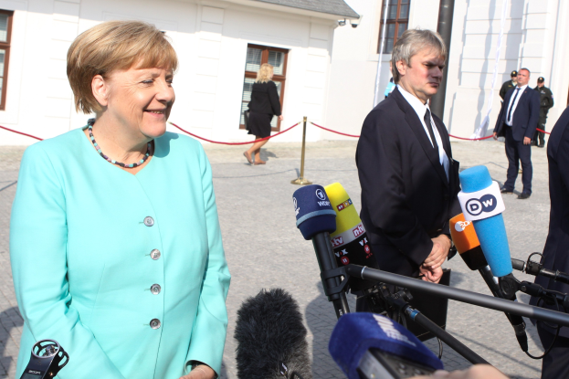 Bundeskanzlerin Angela Merkel spricht vor einem Gebäude mit Fenstern zu Reportern im Freien, umgeben von Menschen und Medienpersonal mit Mikrofonen und Ständern, eine Absperrleine ist sichtbar, der Himmel ist bewölkt.