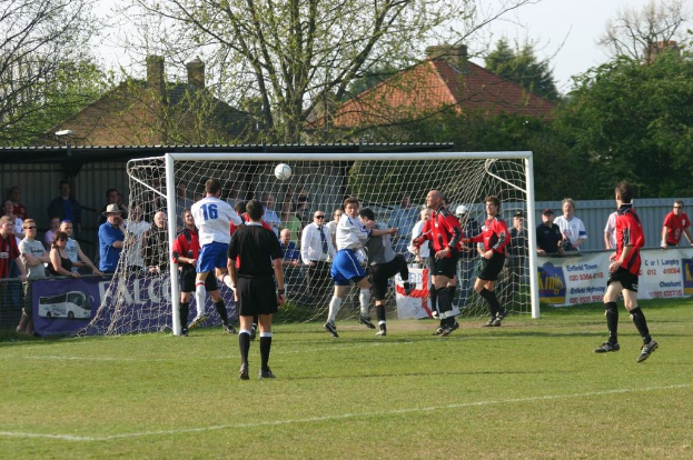 Fußballspieler spielen auf einem Feld mit einem Tor im Hintergrund, während Zuschauer zuschauen, mit Bäumen und Häusern im Hintergrund.