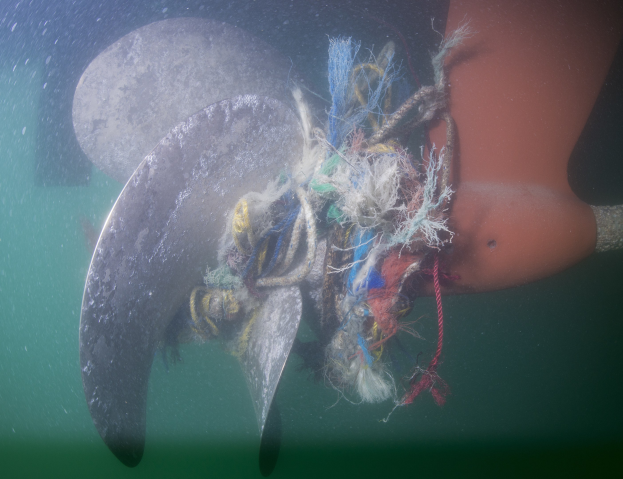 Ein Manta, der im Ozean schwimmt und an dessen Rücken ein großes Propellerblatt befestigt ist, umgeben von farbigen Fäden und Gegenständen.