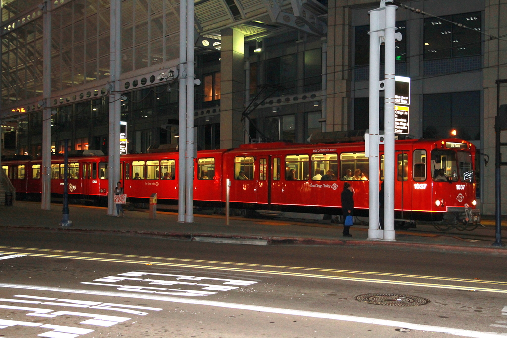 Eine Gruppe von Menschen sitzt in einem roten Zug, mit Pfählen, Schildern und Lichtern draußen.
