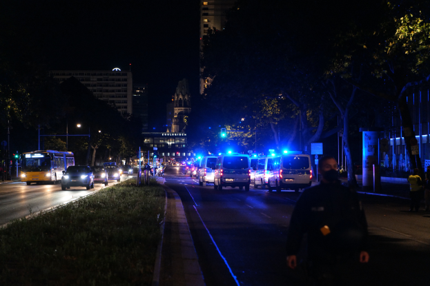 Eine nächtliche Stadtstraße mit Polizei- und Busfahrzeugen, Menschen auf dem Gehweg, Gras auf der linken Seite, Bäumen und Gebäuden im Hintergrund und Laternen, die die Straße beleuchten.