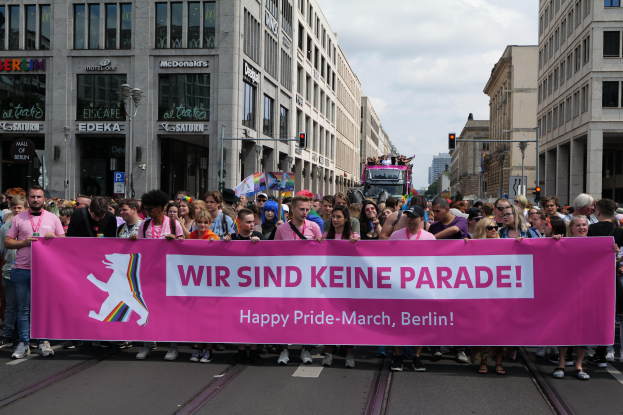 Eine Gruppe von Menschen marschiert auf einer Straße in Berlin, Deutschland, mit einem pinken 'Happy Pride March'-Schild, mit Gebäuden, Laternenmasten und Verkehrszeichen an der Straße.