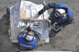 Zwei Männer in blauen Uniformen und Helmen arbeiten an einem großen Drohnenmodell und halten verschiedene Gegenstände, während eine Plastikabdeckung auf dem Boden liegt.