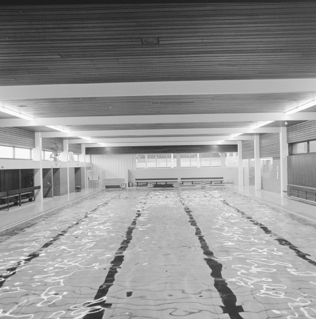 Ein Schwarz-Weiß-Foto eines großen Innenpools mit Wasser auf dem Boden, Bänken, Säulen, Wänden, Fenstern und Deckenlampen in einem geräumigen Gebäude.