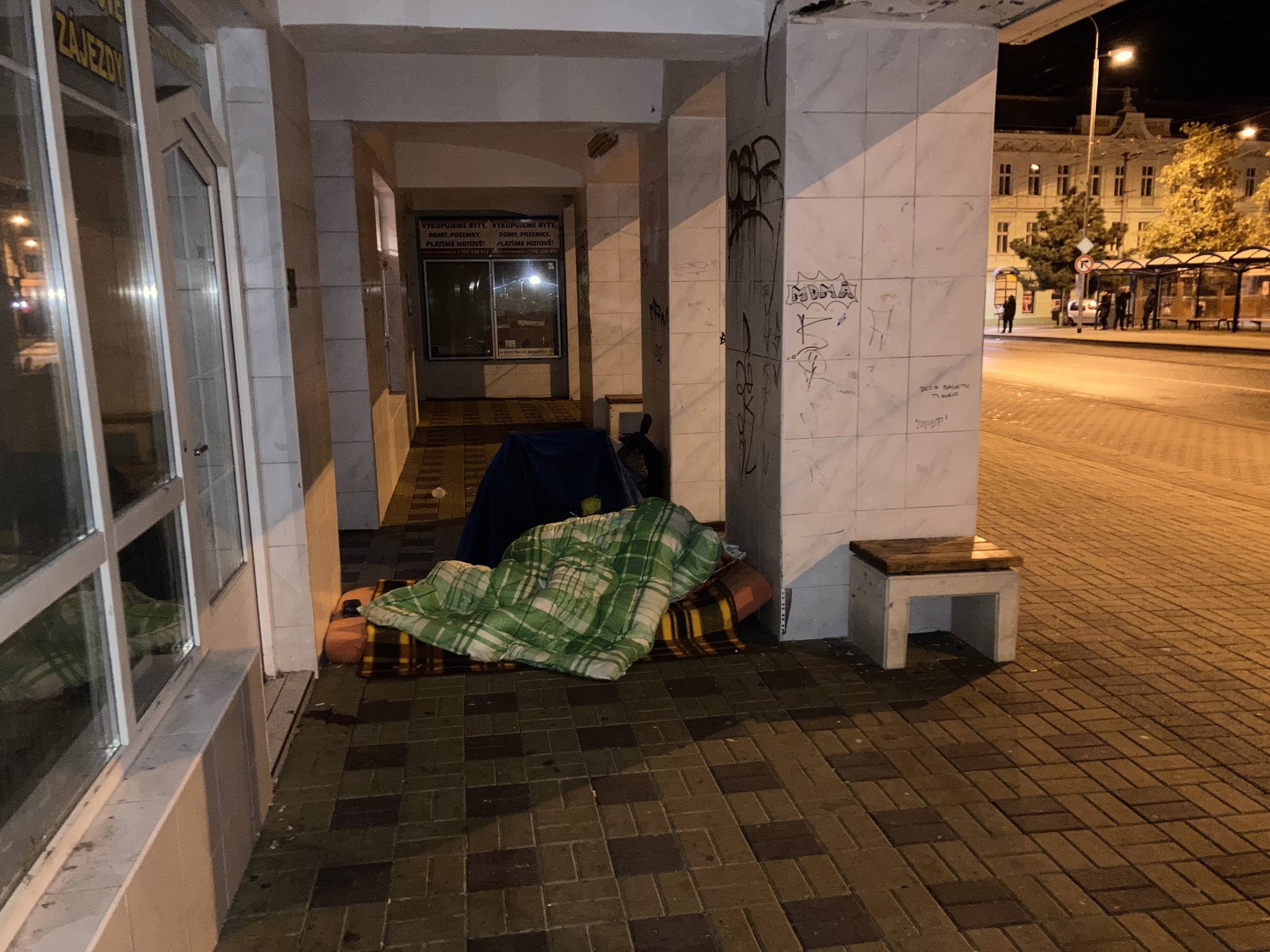 Eine obdachlose Person, die auf einem Bürgersteig bei Nacht schläft, mit einer Decke bedeckt, mit einem Hocker in der Nähe, vor einem Gebäude mit einer Schautafel, einem Straßenschild, Bäumen, Fahrzeugen, Passanten und anderen Gebäuden unter einem bewölkten Himmel.