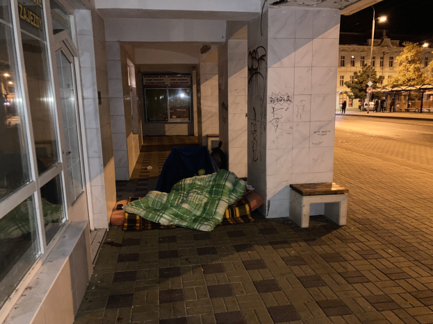 Eine obdachlose Person, die auf einem Bürgersteig bei Nacht schläft, mit einer Decke bedeckt, mit einem Hocker in der Nähe, vor einem Gebäude mit einer Schautafel, einem Straßenschild, Bäumen, Fahrzeugen, Passanten und anderen Gebäuden unter einem bewölkten Himmel.