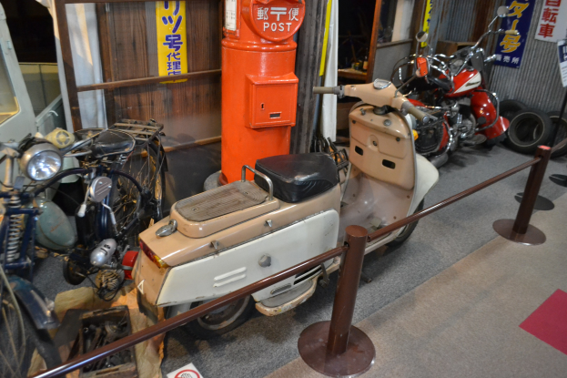 Ein Raum voller verschiedener Motorräder, darunter ein Roller, die nebeneinander geparkt sind, mit einer Stange und einem Seilgeländer im Vordergrund, Bannern mit Text an den Wänden im Hintergrund und Reifen, die herumliegen.