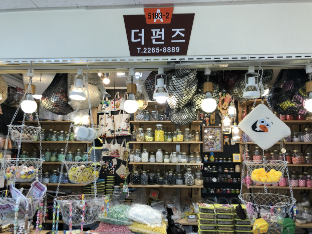 Ein unordentlicher Laden in Seoul zeigt verschiedene Artikel wie Körbe, Lampen, Flaschen und Spielzeuge auf Regalen, mit einer Tafel oben, die ihn als einen der besten koreanischen Läden kennzeichnet.