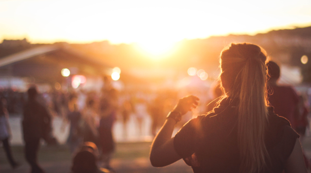 Frau vor einer Menge auf einem Musikfestival bei Sonnenuntergang, mit einem Himmel in warmen Orangetönen, Pink und Lila.