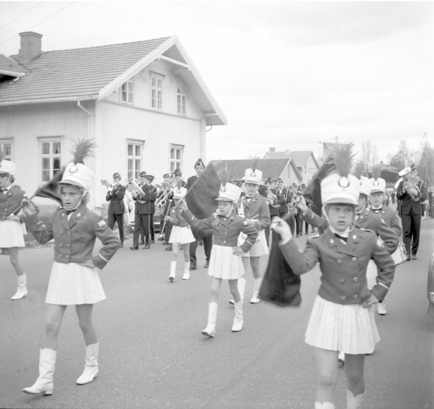 Eine Gruppe junger Mädchen in Uniform marschiert auf einer Straße, trägt Mützen und hält Fahnen, mit Gebäuden, Bäumen und einem bewölkten Himmel im Hintergrund und einem Fahrzeug auf der rechten Seite.