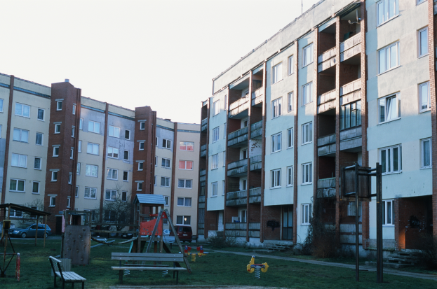 Ein Wohnhaus mit einem Spielplatz davor, mit Rasen, Bänken, Schaukeln und anderen Gegenständen, mit Fahrzeugen, Bäumen und einem klaren blauen Himmel im Hintergrund.