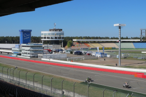Eine Gruppe von Menschen auf Motorrädern auf einer Rennstrecke umgeben von einem Zaun, Fahrzeugen, einem Gebäude, Schildern, einem Stadion und Bäumen unter einem bewölkten Himmel.