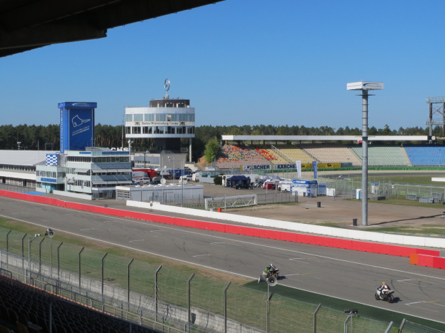 Eine Gruppe von Menschen auf Motorrädern auf einer Rennstrecke umgeben von einem Zaun, Fahrzeugen, einem Gebäude, Schildern, einem Stadion und Bäumen unter einem bewölkten Himmel.