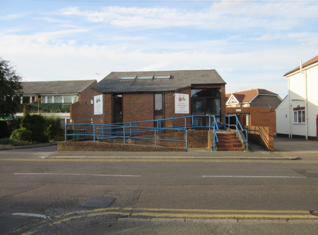 Ein Schulgebäude mit einem blauen Geländer davor, umgeben von Häusern, Treppen mit Geländern, Informationsschildern, Pflanzen, Bäumen und einem bewölkten Himmel.