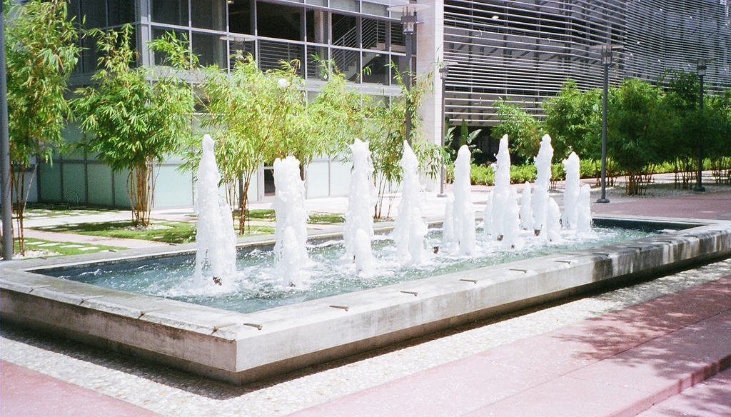 Eine Wasserfontäne mit Pflanzen, Straßenlaternen und Gras im Vordergrund und Gebäuden im Hintergrund, mit Stufen unten.